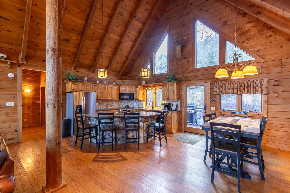 This image captures a spacious, open-plan kitchen and dining area, featuring wooden beams, large windows that frame natural views, a central kitchen island, and a generous dining table. Natural light fills the space, and the setup invites communal cooking and gathering.
