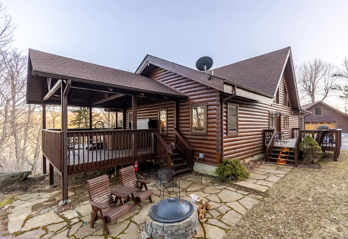 The image showcases a charming log cabin nestled in the woods, featuring a spacious deck with inviting outdoor seating, a cozy fire pit, and space for al fresco dining. The setting is tranquil and perfect for relaxation, with hints of sunset light creating a warm, welcoming atmosphere.