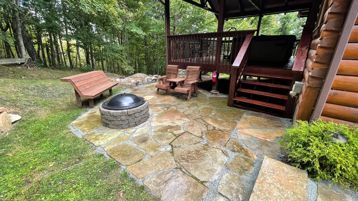 A cozy stone patio nestled amidst lush greenery, featuring inviting seating around a central fire pit. A stone pathway winds through the yard, suggesting easy access to both nature and social gatherings.