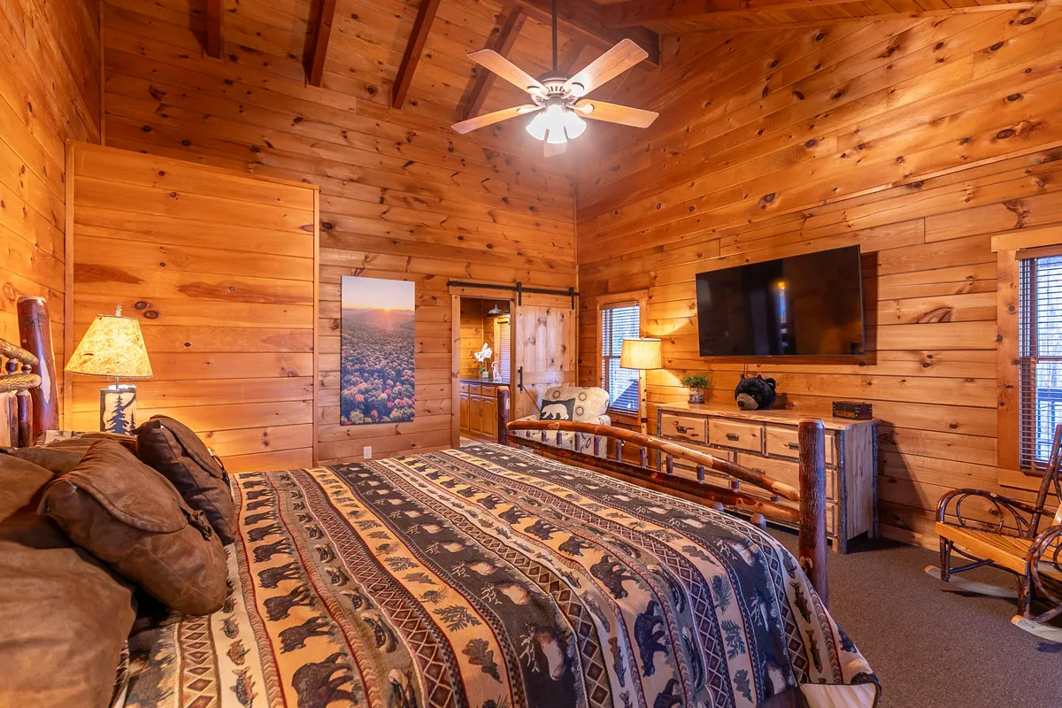A spacious bedroom with a large bed featuring a patterned blanket, a flat-screen TV, decorative lamp, and wooden furniture. The room exudes warmth with its wooden ceiling and cozy design elements, creating an inviting space for relaxation and quiet moments.