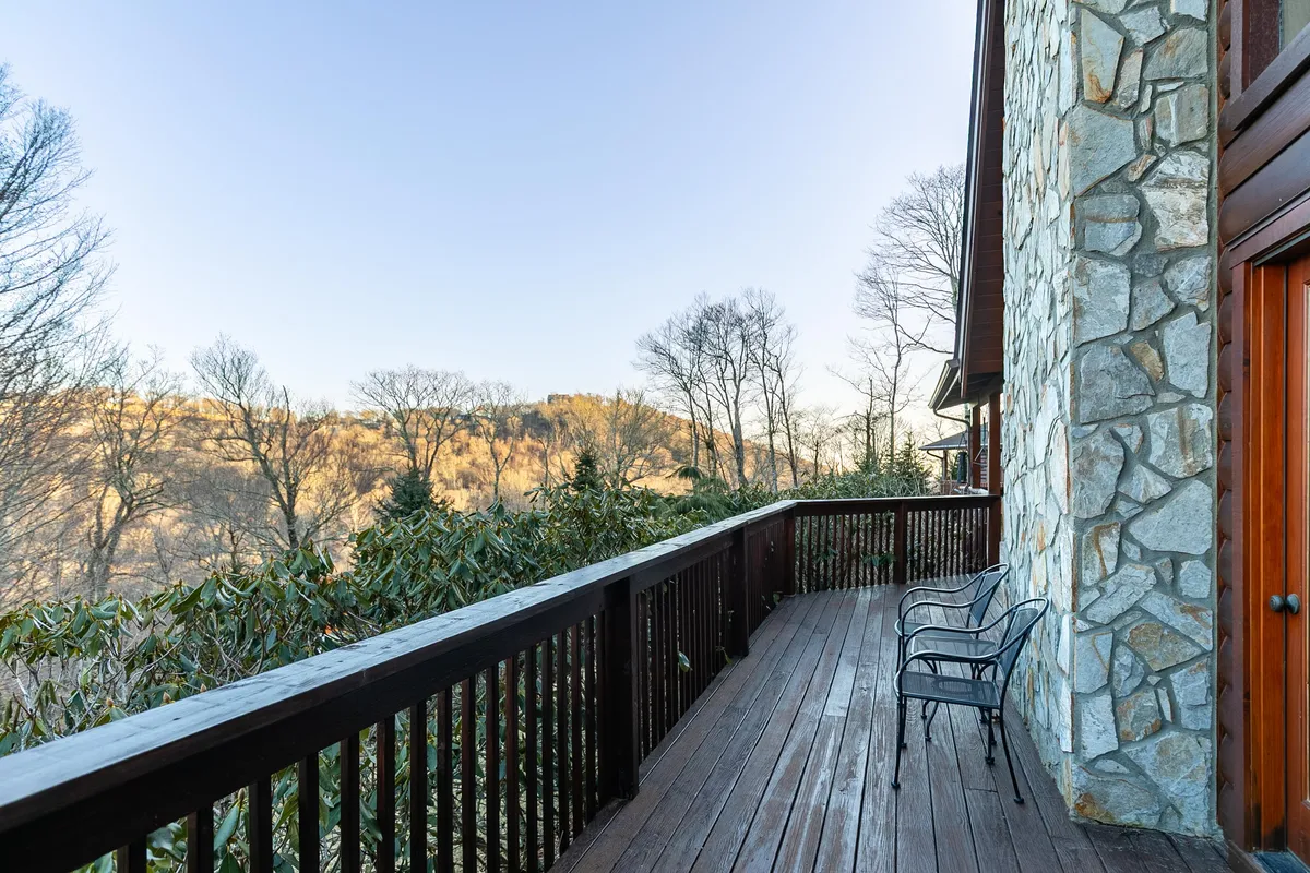 This image presents a tranquil wooden deck with two metal chairs overlooking rolling hills and distant mountains. The setting is serene, with soft natural light suggesting either early morning or twilight. The view is expansive, capturing the peaceful seclusion and natural beauty ideal for couples seeking a private retreat.