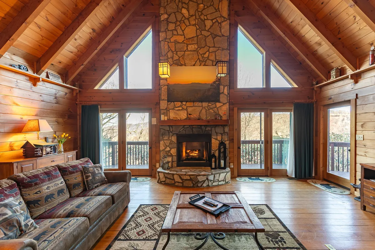 This image showcases a warm, rustic living room featuring a grand floor-to-ceiling stone fireplace, comfortable sofa seating, large windows, exposed wooden beams, and a cozy coffee table. The fireplace is crackling, and the ample plush seating is arranged for conversation or relaxation, with natural light streaming in from the windows.