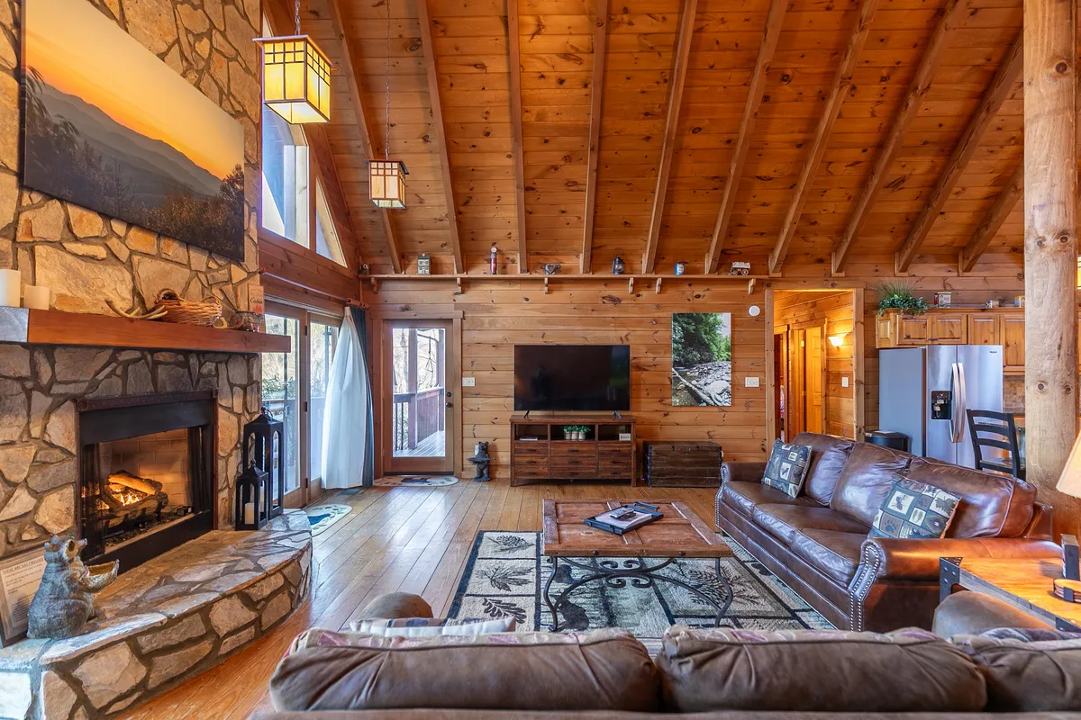 A beautifully appointed living area with a striking stone fireplace, leather sofas, wooden ceiling beams, and large windows that offer peeks of the outdoor scenery. The seating is arranged for socializing, and the decor highlights a warm, welcoming atmosphere ideal for gatherings or quiet moments by the fire.