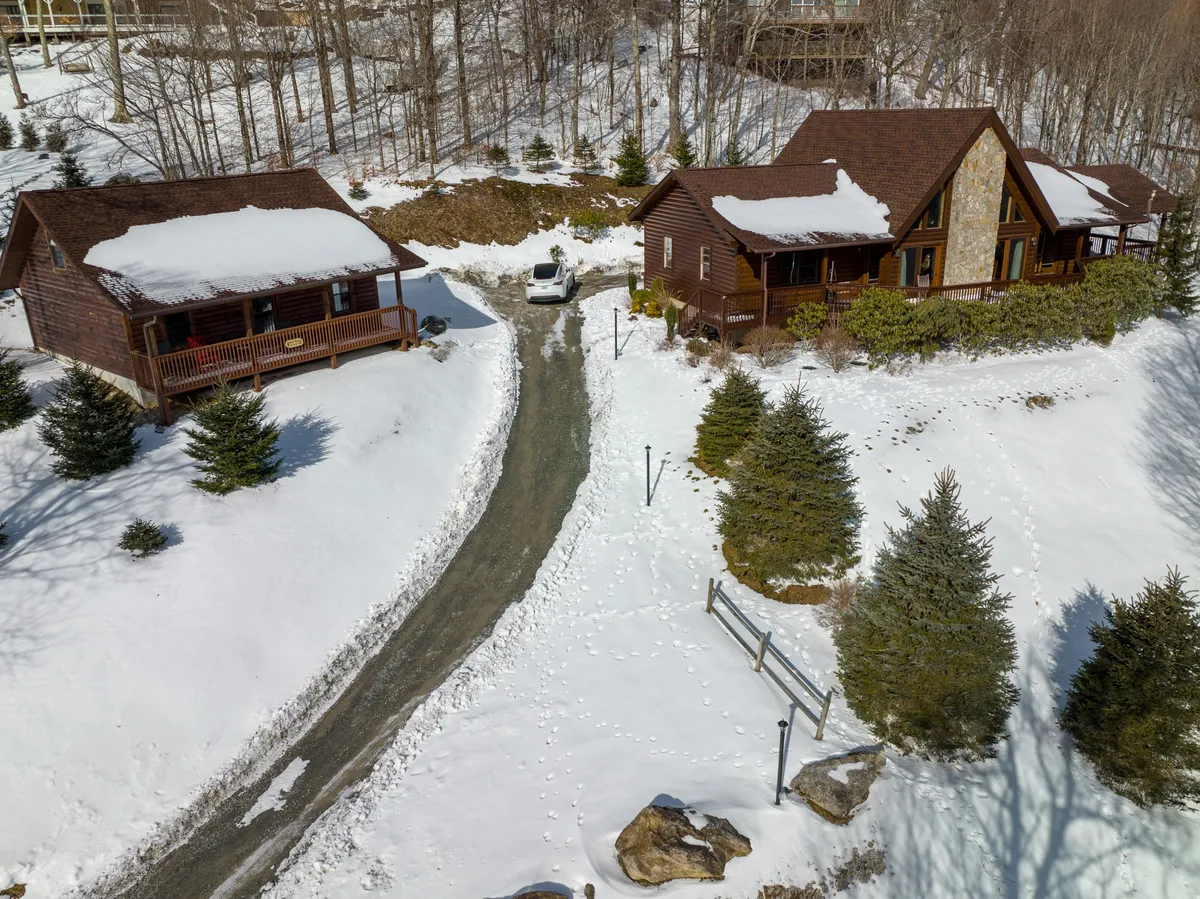Two charming log cabins nestled in a snowy landscape with a gravel driveway and evergreen trees. The setting feels both cozy and adventurous, perfect for families or groups after a day on the slopes.