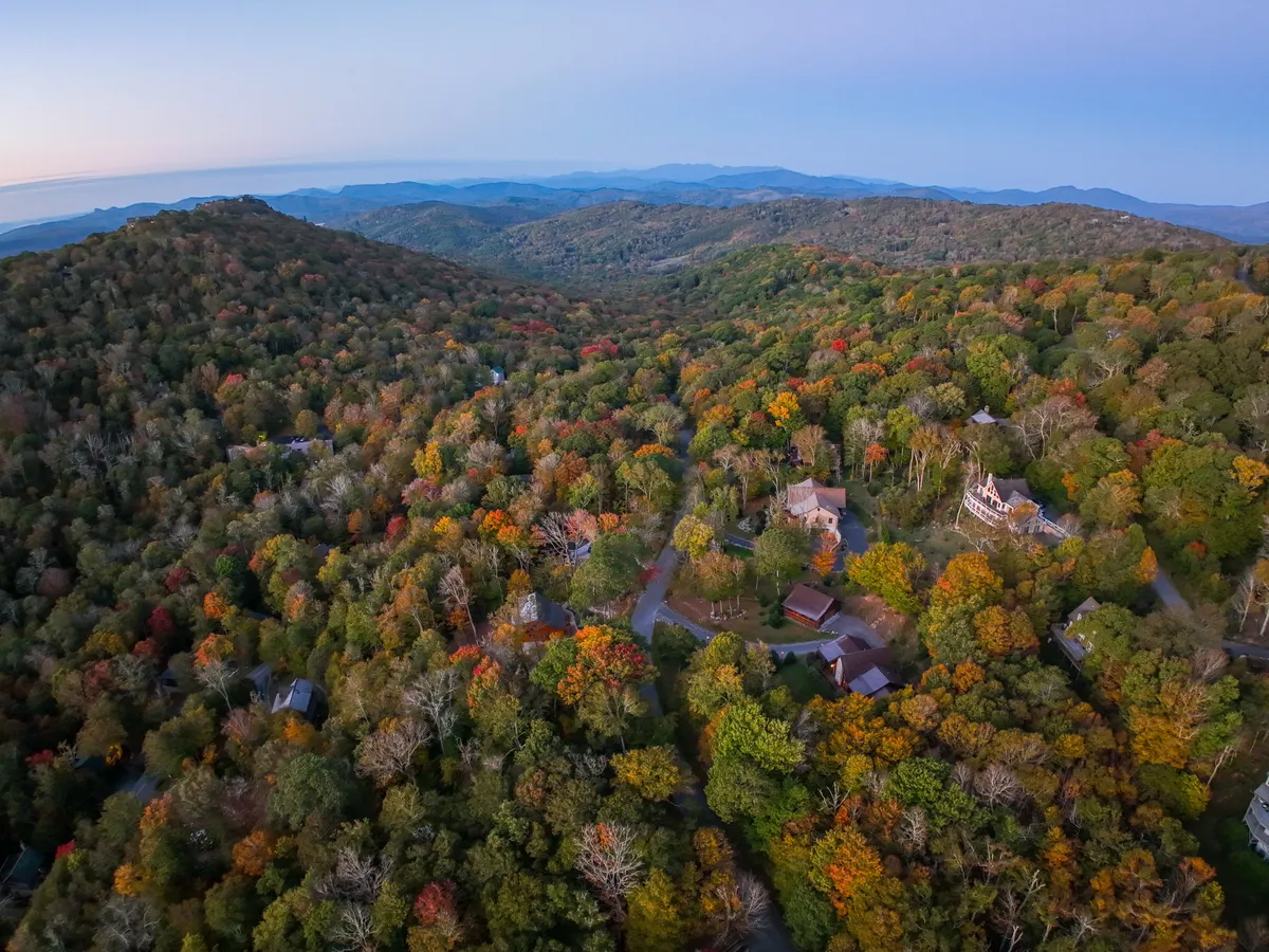 A sweeping view of rolling hills blanketed in vibrant, colorful foliage, with distant mountains rising against the horizon. Scattered homes dot the landscape, and the scene radiates tranquility and the boundless beauty of nature, ideal for outdoor activities like hiking, nature walks, and photography.