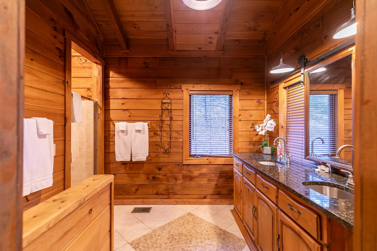 A well-appointed hallway bath with granite countertops, wooden cabinetry, large mirrors, and a window with blinds. The space feels spacious and spa-like—perfect for morning routines or unwinding after the day’s activities.