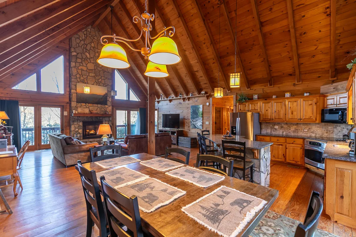 The photo highlights a warm, inviting dining area adjacent to a modern kitchen, accentuated by wooden beams, a central stone fireplace, large windows, and a robust wooden dining table that seats a group. The scene suggests comfort and togetherness, with ample space for family meals and socializing.