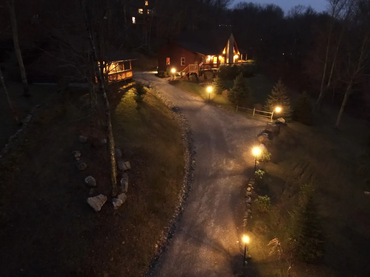 An exterior shot taken at dusk, showing the warmly illuminated house along a winding gravel path, surrounded by trees. Pathway lights create a cozy, welcoming glow, and the tranquil night sky hints at opportunities for stargazing and outdoor relaxation.
