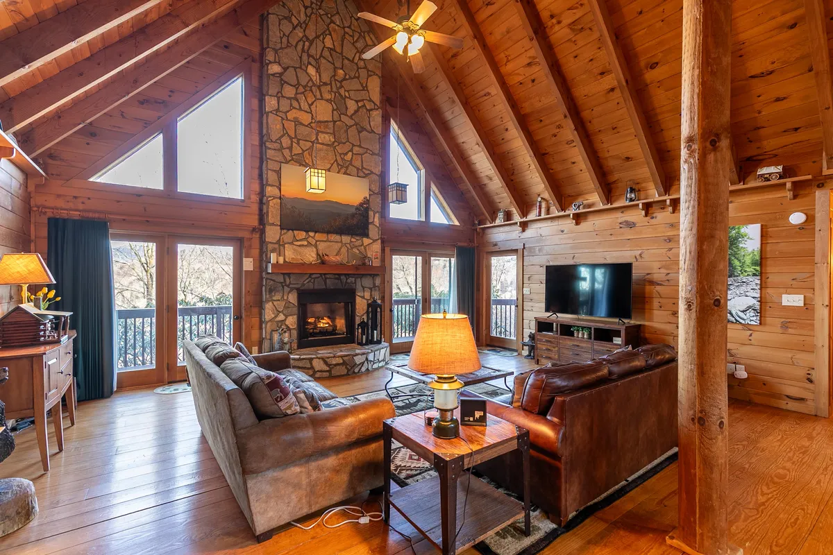 A warmly lit living room featuring a stone fireplace, large windows with views, wooden beams, and a plush leather sofa. The inviting space is arranged for cozy relaxation, perfect for intimate nights in by the fire.