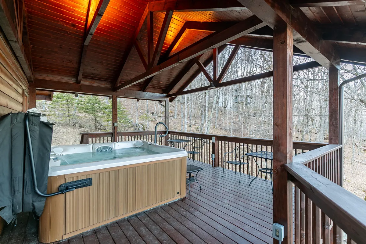 This exterior photo shows a hot tub on a spacious wooden deck surrounded by tall trees. The hot tub is the focal point but the image also highlights the generous deck space for relaxing or socializing.