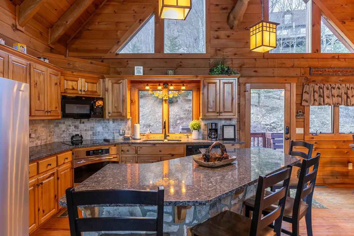 A close-up of a granite kitchen countertop with wooden cabinetry, large windows bringing in daylight, and elegant hanging light fixtures. This perspective highlights the practical and stylish features of the kitchen, underscoring its readiness for culinary adventures.