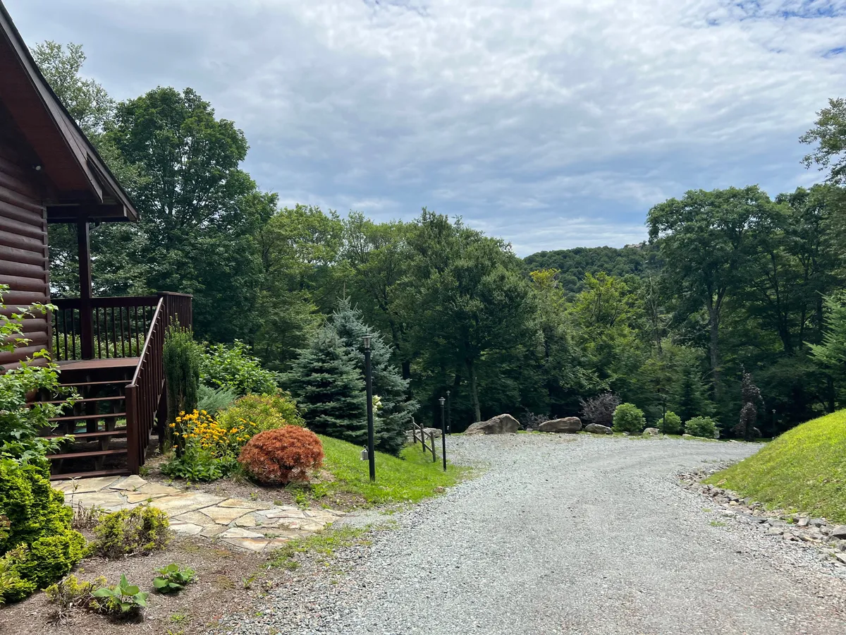 A log cabin nestled in lush greenery with a welcoming porch, gravel driveway, and blooming flower beds. The scene exudes tranquility and showcases the outdoor areas perfect for nature walks or relaxing with your dog.